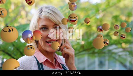 Donna sorridente che indossa un grembiule verde che riceve chiamate sullo smartphone in giardino, con sfere emoji fluttuanti Foto Stock