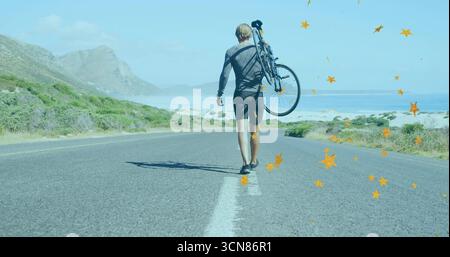 Ciclista maschile che cammina portando la bicicletta a spalla lungo l'autostrada costiera, indossando un abbigliamento da ciclismo Foto Stock