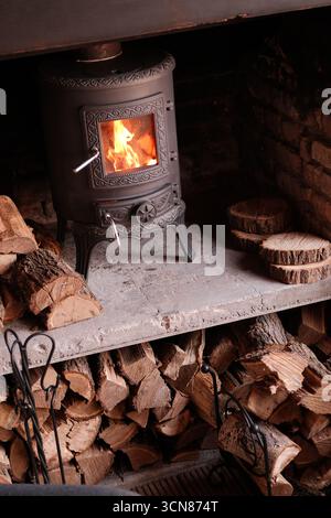 Un'accogliente stufa a legna che emette un fuoco caldo con logge ben impilate nelle vicinanze per migliorare il comfort Foto Stock