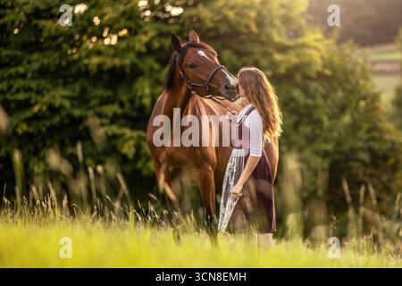 Ritratto romantico di una giovane donna in abito tradizionale con il suo cavallo trotter della baia all'aperto Foto Stock