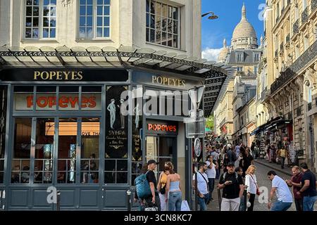 Parigi, Francia. 8 settembre 2025. Una vivace scena di strada a Parigi con un fast-food Popeyes al piano terra di un edificio storico. I pedoni camminano lungo il marciapiede mentre una stretta strada si estende verso la cupola illuminata dell'area Sacré-Cœur visibile sullo sfondo l'8 settembre 2025. (Foto di Samuel Rigelhaupt/Sipa USA) credito: SIPA USA/Alamy Live News Foto Stock