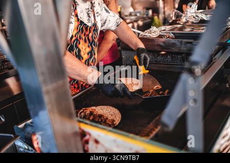 Lo chef indossa guanti e un grembiule a fiamma che prepara deliziose quesadillas con funghi e altri ingredienti su una piastra calda in una vivace st Foto Stock