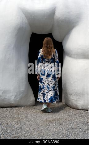 Una persona dai capelli lunghi con un abito floreale blu e bianco cammina verso un ingresso scuro dell'edificio incorniciato da due grandi architetture bianche di forma organica Foto Stock
