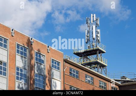 Una torre di telefoni cellulari con più antenne si trova in cima a un edificio di mattoni rossi contro un cielo blu luminoso, evidenziando le moderne infrastrutture di telecomunicazione Foto Stock