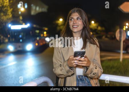 Giovane donna sorridente mentre controlla il suo telefono cellulare alla fermata dell'autobus della città di notte, anticipando il trasporto o incontrando gli amici in un ambiente urbano Foto Stock