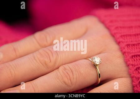 La mano di una donna tiene in mano un anello con un diamante dentro. L'anello è d'oro e la donna indossa un maglione rosa Foto Stock