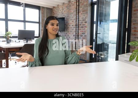 Donna che scrive sulla tastiera al tavolo in un ufficio moderno e indossa una camicetta verde vicino al monitor del computer Foto Stock