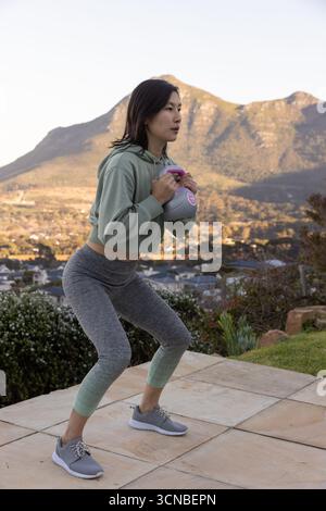 Donna asiatica coreana che esegue squat ponderato in abbigliamento sportivo su una terrazza piastrellata con kettlebell Foto Stock