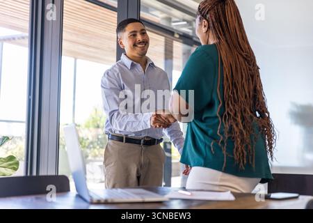 Diversi colleghi che stringono la mano sul tavolo da conferenza in legno in ufficio con computer portatile e documenti Foto Stock