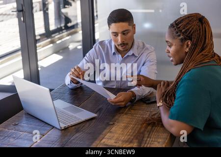 Colleghi diversi che indossano abiti da lavoro che rivedono documenti con penna e laptop in ufficio Foto Stock