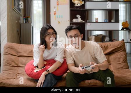 la coppia si siede su un comodo divano del soggiorno e gioca a un videogame. L'uomo mostra frustrazione con il controller, mentre la donna sembra sorpresa Foto Stock