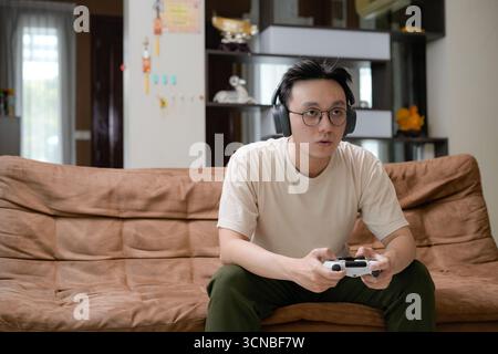 Un giovane uomo siede su un divano marrone con cuffie di grandi dimensioni e gioca su un controller di gioco Foto Stock