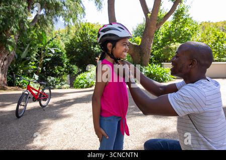 Padre e figlia afroamericani si inginocchiano sul vialetto che fissa la cinghia del casco accanto alla bicicletta rossa Foto Stock