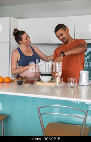 Coppia che indossa un abbigliamento sportivo e una maglietta dietro il bancone della cucina che mescola frutti di bosco e arance nel frullatore Foto Stock