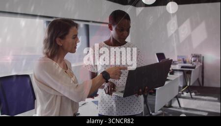 Tenendo un notebook e puntando sullo schermo, due donne collaborano in un ufficio open space, con monitor Foto Stock