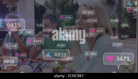 Amici seduti al tavolo da picnic nel parco, mangiando panini e hamburger con sovrapposizioni fluttuanti di icone Foto Stock