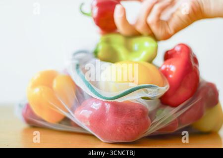 Peperoni multicolore in un sacchetto riutilizzabile ecologico e una mano che tiene in mano una paprika rossa sullo sfondo. Cultura da fattoria a tavola e dieta vegetariana Foto Stock