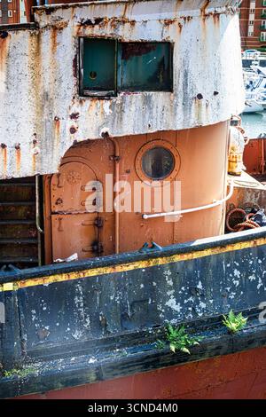 Un primo piano di una nave in decadenza mostra la vernice sbucciata, la ruggine profonda e le piccole piante che iniziano a recuperare i bordi esterni. Foto Stock