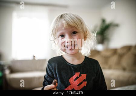 Una gioiosa bambina di 5 anni celebra Halloween nel suo accogliente salotto. Vestita con un costume divertente, sorride brillantemente mentre sorseggia una sorpresa, circondata Foto Stock