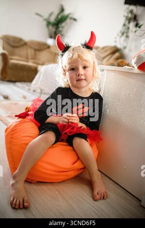 Una bambina di 5 anni vestita con un costume di Halloween si rilassa su un sacchetto di fagioli arancioni. Ha corna del diavolo giocose e un'espressione felice mentre ama la f Foto Stock