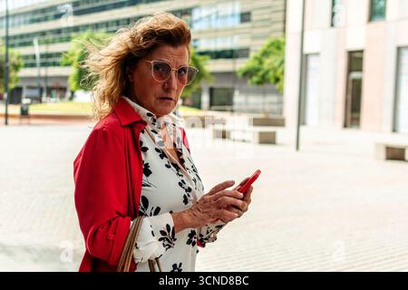 Donna dirigente senior sicura che indossa occhiali da sole e tiene in mano uno smartphone mentre cammina in un ambiente urbano moderno, mostrando la sua professionalità Foto Stock