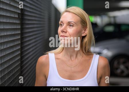 Ritratto di una donna bionda sicura di sé accanto a una recinzione metallica in un ambiente urbano, con una canotta bianca e una collana dorata. Foto Stock
