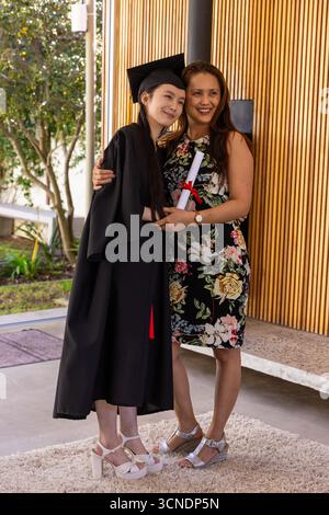 Madre-figlia che abbraccia e sorride sul patio indossando cappello e abito neri, tenendo in mano il rotolo del diploma Foto Stock