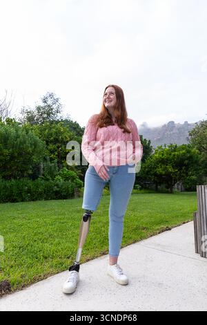 Donna in piedi su un sentiero pavimentato nel parco che indossa un maglione rosa e jeans blu che mostra un arto protesico Foto Stock