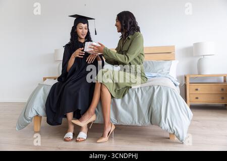 Mamma e figlia adolescente diverse si scambiano una piccola confezione regalo indossando un abito da cerimonia in camera da letto Foto Stock