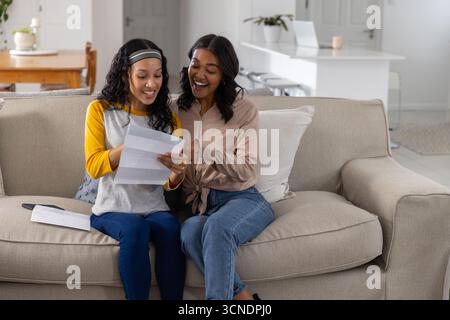 Mamma e figlia adolescente diverse sedute sul divano leggono una lettera con busta nel soggiorno Foto Stock