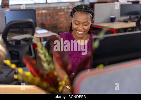 Donna afroamericana che indossa la parte superiore magenta, sorridente, che digita la tastiera in ufficio con il monitor Foto Stock