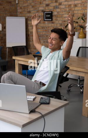 Uomo asiatico che si allunga le braccia sopra la testa alla scrivania in ufficio con laptop smartphone lavagna a fogli mobili Foto Stock