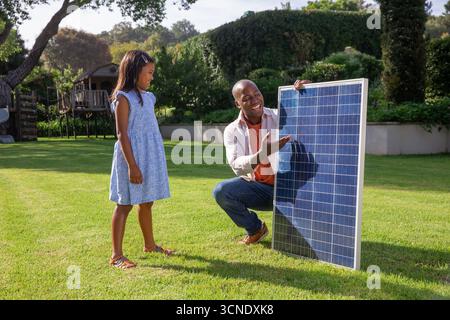 Padre e figlia afroamericani che esaminano un grande pannello solare nel cortile di periferia nella giornata di sole Foto Stock