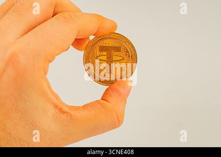 Una mano regge una moneta Tether dorata con il suo simbolo, su un semplice sfondo questa immagine potrebbe essere utilizzata per l'educazione alla criptovaluta, il marketing, o Foto Stock