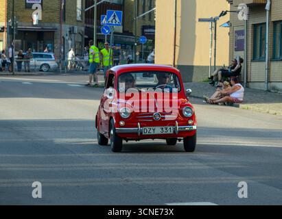 Una piccola e classica auto rossa si muove lungo una strada trafficata in una giornata di sole. Gli spettatori, comprese famiglie e amici, si riuniscono nelle vicinanze per godersi l'estate ad atmos Foto Stock
