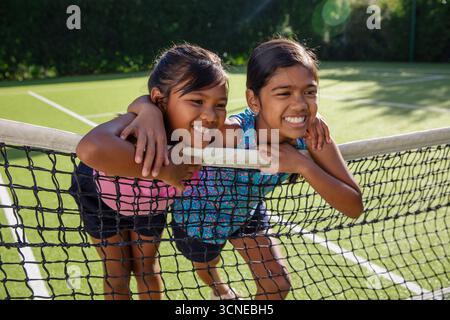 Bambine amiche che si appoggiano su una rete da tennis su un campo da tennis in erba indossando abbigliamento sportivo Foto Stock