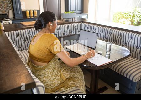Donna dell'asia meridionale che indossa sari gialli senape e scrive su un notebook al tavolo con un computer portatile Foto Stock