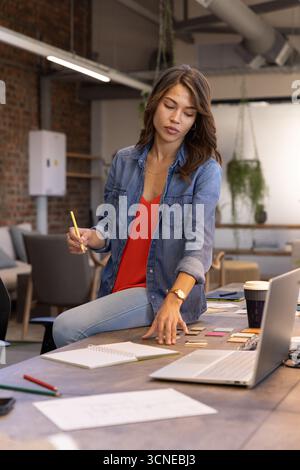 Donna designer seduta sulla scrivania in un moderno ufficio di coworking che rivede i campioni a colori con un computer portatile Foto Stock