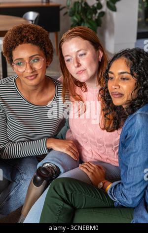 Diverse amiche di sesso femminile sedute sul divano di casa indossano occhiali da vista, con protesi alle gambe Foto Stock