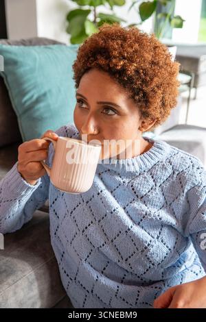 Donna afroamericana che sorseggia una tazza bianca sul divano nel soggiorno con cuscino di tè Foto Stock