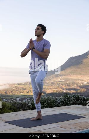 Uomo afroamericano in abbigliamento sportivo in piedi su un tappetino da yoga su un patio piastrellato che si affaccia sulla valle Foto Stock