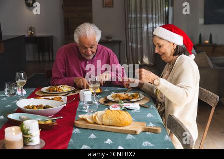 Coppia senior che indossa il cappello di Babbo Natale tagliando pollo e verdure nella sala da pranzo con bicchieri da vino Foto Stock
