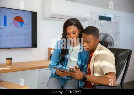 Colleghi afroamericani in abbigliamento da lavoro che collaborano con un tablet alla scrivania dell'ufficio con un monitor Foto Stock
