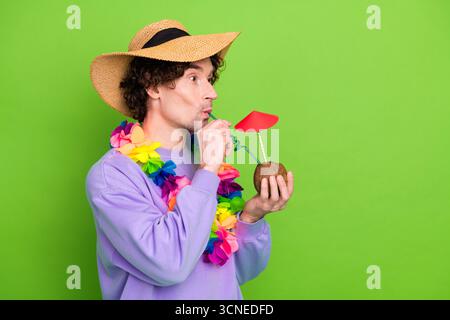 Giovane uomo in abbigliamento informale che si gode una bevanda tropicale su un vivace sfondo verde, indossa accessori colorati e un cappello di paglia. Foto Stock