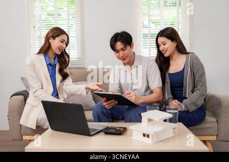Un agente immobiliare asiatico professionale consulta i clienti a casa. Discutono di investimenti immobiliari e finalizzano l'accordo di acquisto nel comfort moderno. Foto Stock