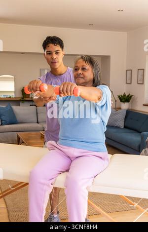 Cliente donna anziana e addestratrice che solleva manubri arancioni sulla panchina nel soggiorno, postura di guida Foto Stock