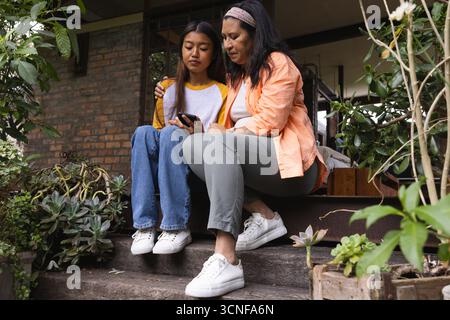 Madre ispanica e figlia asiatica adolescente seduta sui gradini del portico che condividono lo smartphone tra le piante Foto Stock