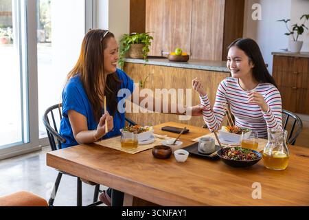 Mamma e figlia sedute a tavola in cucina a condividere l'insalata con le bacchette e a chiacchierare. Foto Stock