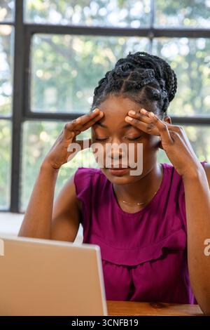 Donna afro-americana che preme le tempie, chiude gli occhi alla scrivania con un laptop accanto alla finestra Foto Stock