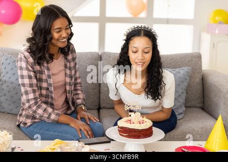 Mamma e figlia adolescente diverse festeggiano il diciottesimo compleanno sul divano grigio di casa con torta Foto Stock